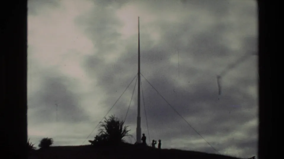 Handheld 8mm view of the Flagstaff Hill pole, guy wires, shrubs, and distant visitors silhouetted against cloud cover.