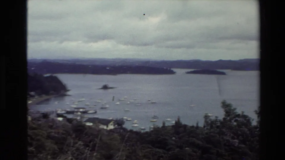 1981 New Zealand home-movie harbor panorama from Flagstaff Hill, with anchored boats, low hills, islands, and foreground vegetation.
