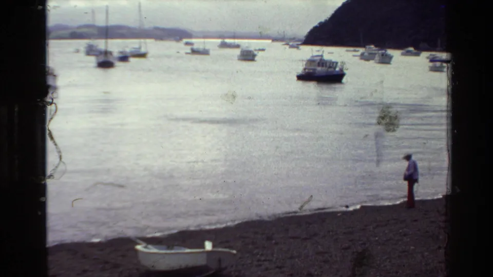 1981 New Zealand shoreline frame with a pebble beach, small rowboat, anchored boats, and one distant person standing near the water.