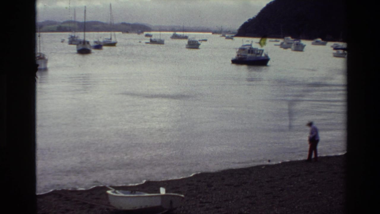 1981 New Zealand 8mm frame showing a shoreline figure, moored harbor boats, distant hills, and a small white rowboat on the pebbles.