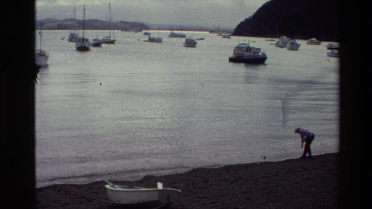 Person bending on a pebble shoreline with moored boats across calm harbor water.