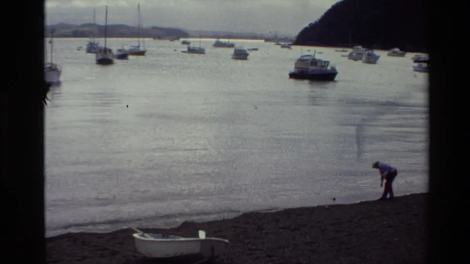 1981 New Zealand home-movie frame with a person bending near the harbor edge, boats at anchor, a rowboat on shore, and overcast light.