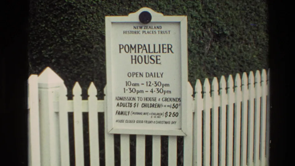1981 New Zealand frame showing a Pompallier House visitor sign with opening hours and admission prices beside a white picket fence.