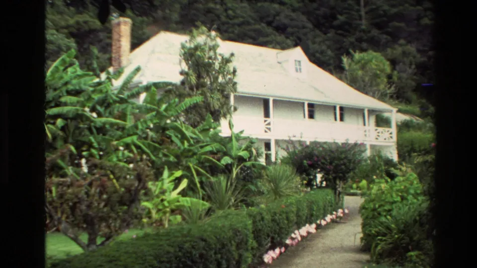 1981 New Zealand 8mm frame with Pompallier House partially framed by banana plants, hedges, and flowering shrubs.