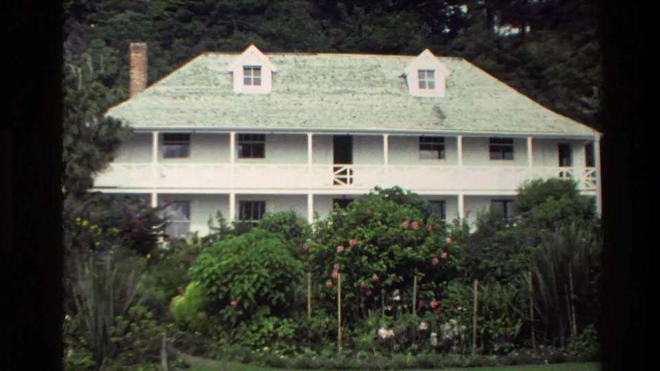 1981 New Zealand 8mm front view of Pompallier House with veranda, dormer windows, chimney, and dense flowering garden beds.