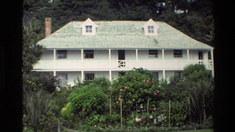 1981 New Zealand 8mm frame showing a white timber house with a pale roof, upper porch, garden shrubs, and trees behind it.