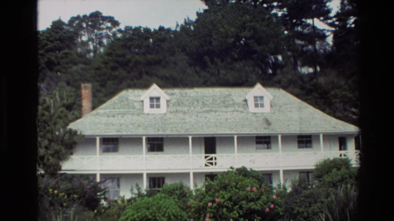 1981 New Zealand exterior view of a white wooden house with dormer windows, veranda railings, dense shrubs, and trees in the background.