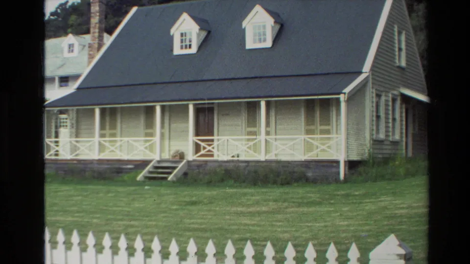 1981 New Zealand frame showing a white cottage with a dark roof, front porch, small steps, mowed lawn, and white picket fence.