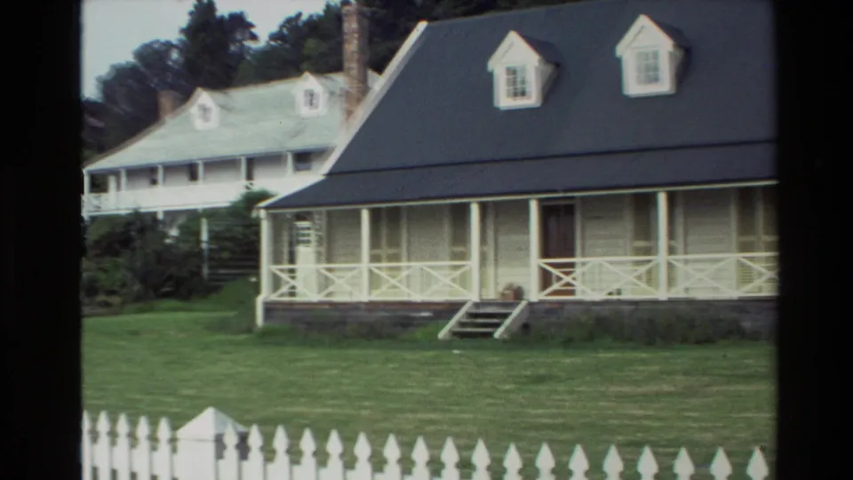 1981 New Zealand 8mm frame with a dark-roofed white cottage, another white house behind it, lawn, porch railings, and picket fence.