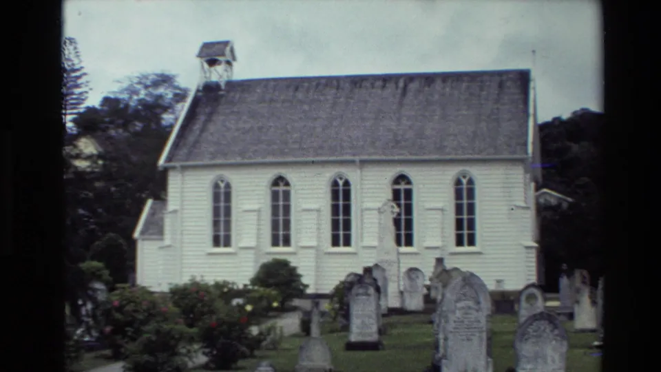 1981 New Zealand frame showing Christ Church Russell's exterior across a churchyard with weathered headstones, shrubs, and arched windows.