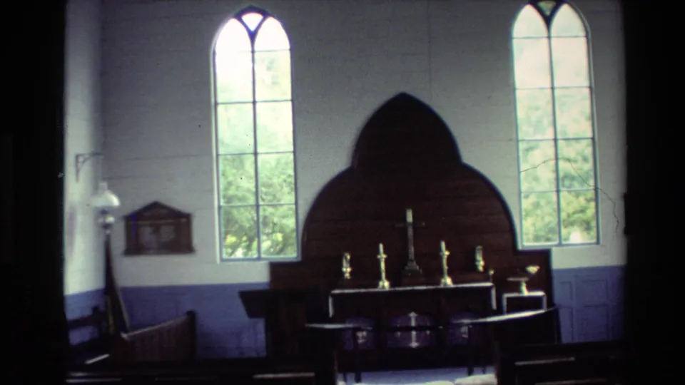 Interior 1981 home-movie view of Christ Church Russell showing a simple altar, candles, wooden pews, and tall arched windows.