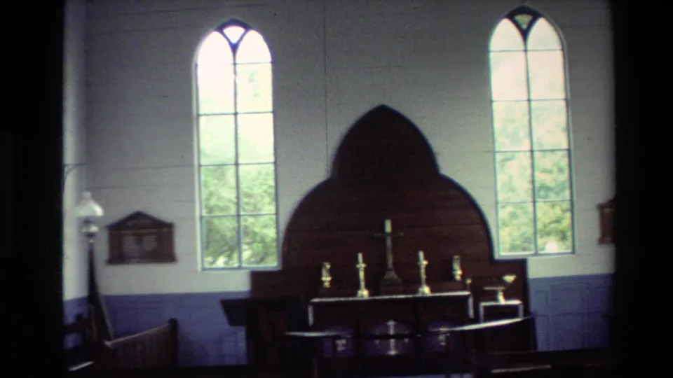 1981 New Zealand 8mm frame of the same church interior from a lower handheld angle, with the altar, cross, candlesticks, pews, and bright windows visible.