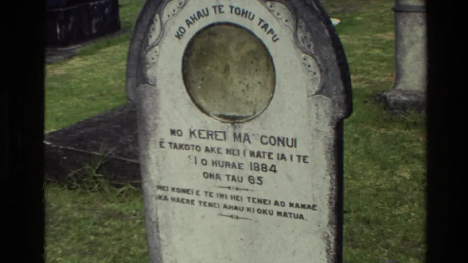 1981 New Zealand cemetery frame showing a rounded headstone with Maori script, circular recess, grass, and another marker nearby.