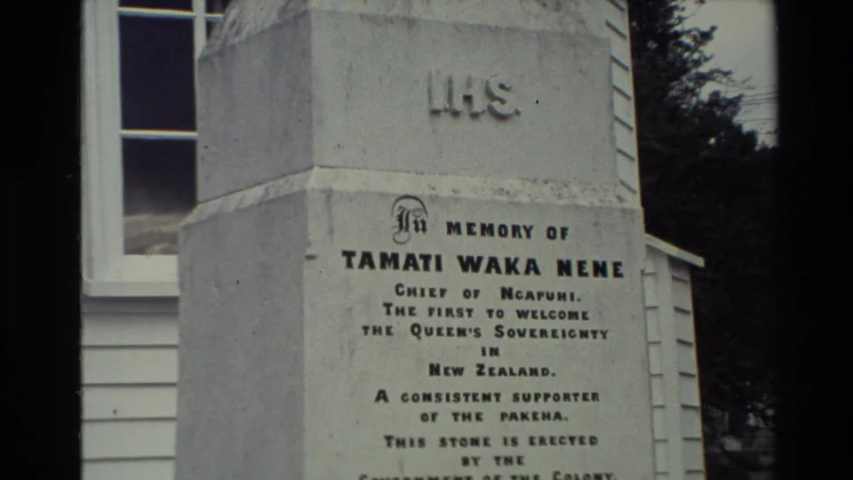 Visitor-shot 8mm view of the Tamati Waka Nene memorial inscription in New Zealand, with white siding and a window behind it.