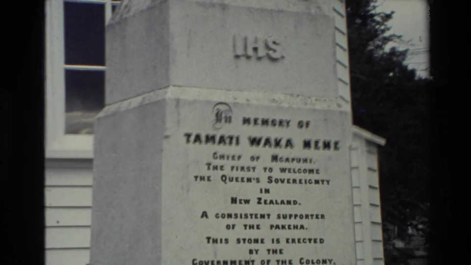 1981 home-movie frame showing the weathered white Tamati Waka Nene memorial with inscription and nearby building details.