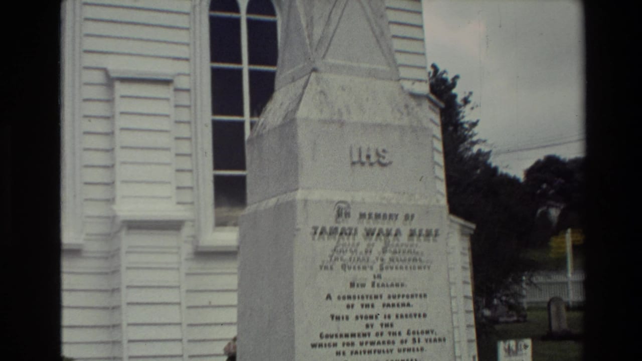 1981 New Zealand home-movie frame showing an inscribed stone memorial beside a white timber church.