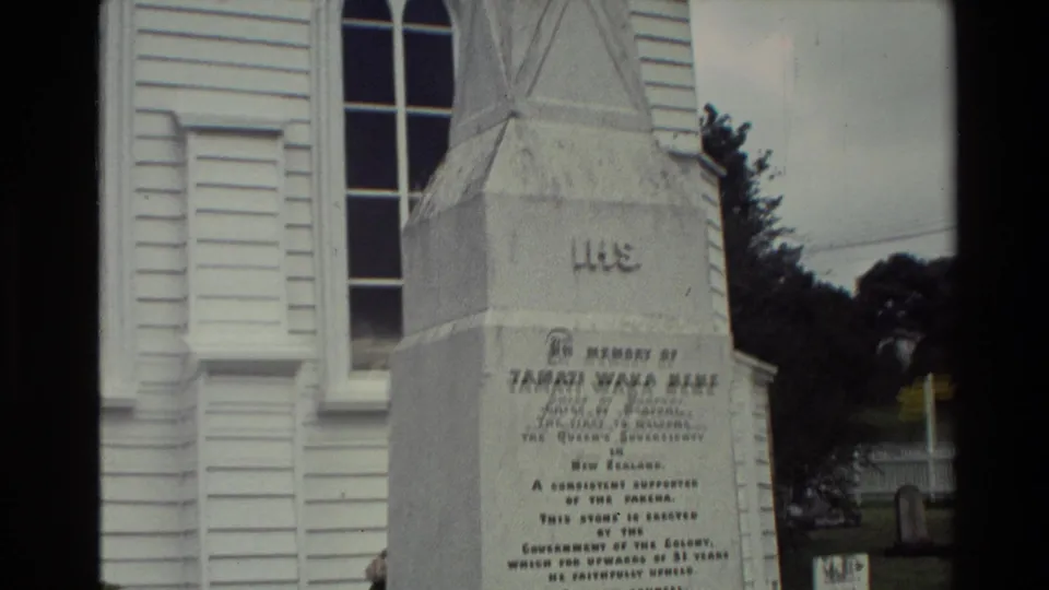 1981 New Zealand home-movie frame showing an inscribed stone memorial beside a white timber church.