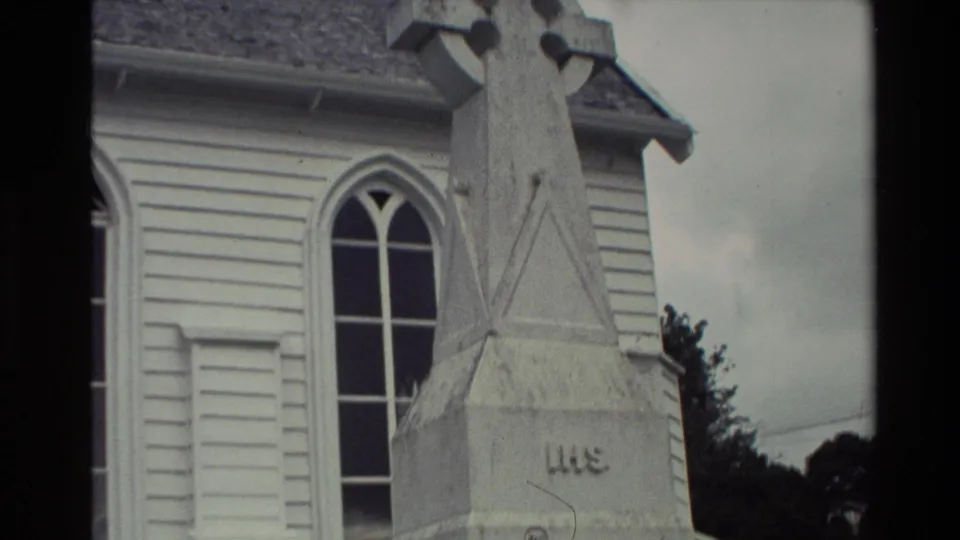 Handheld 1981 New Zealand frame of the upper stone memorial cross near a white church wall and arched window.