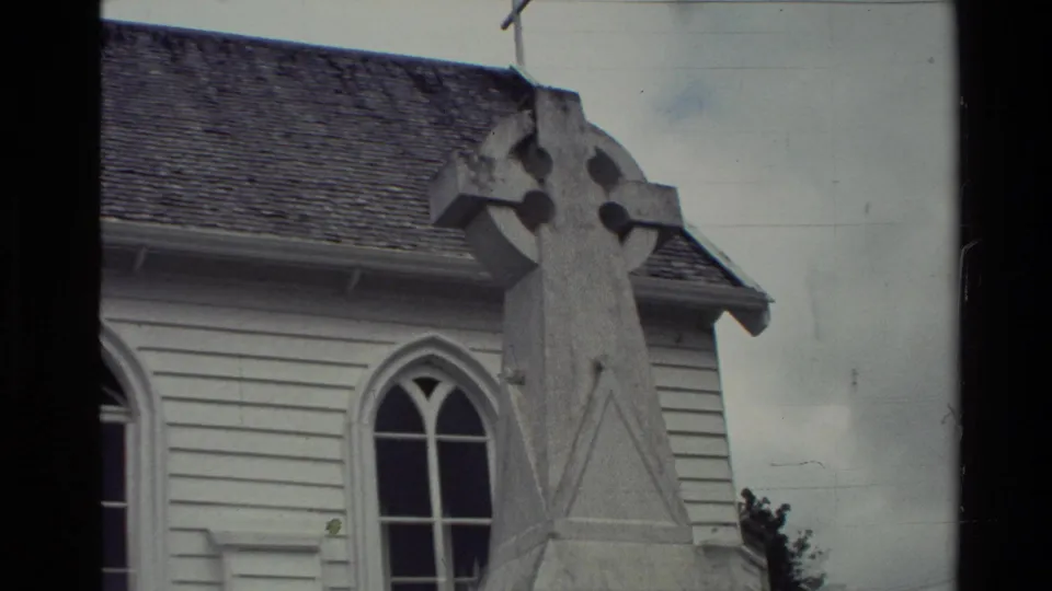A 1981 New Zealand Super 8 frame shows the tall memorial cross beside the church's arched window and shingled roof.