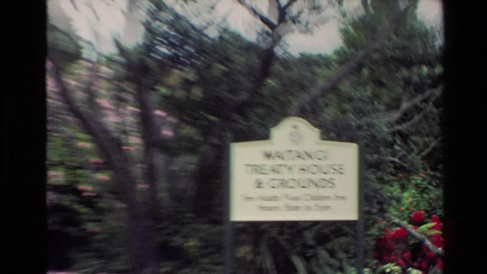 A blurred 1981 New Zealand home-movie frame shows a cemetery sign in greenery with red flowers nearby.