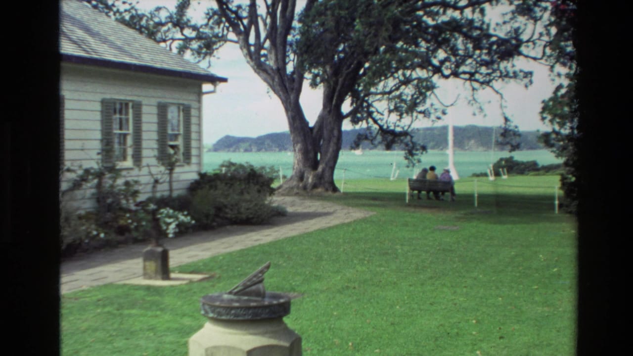1981 New Zealand 8mm frame showing a lawn, white house, large tree, sundial, waterfront, and two distant people seated on a bench.
