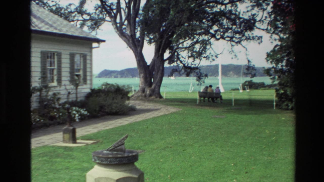 Similar garden view with white house, tree, lawn, sundial, waterfront, and two people on a bench.