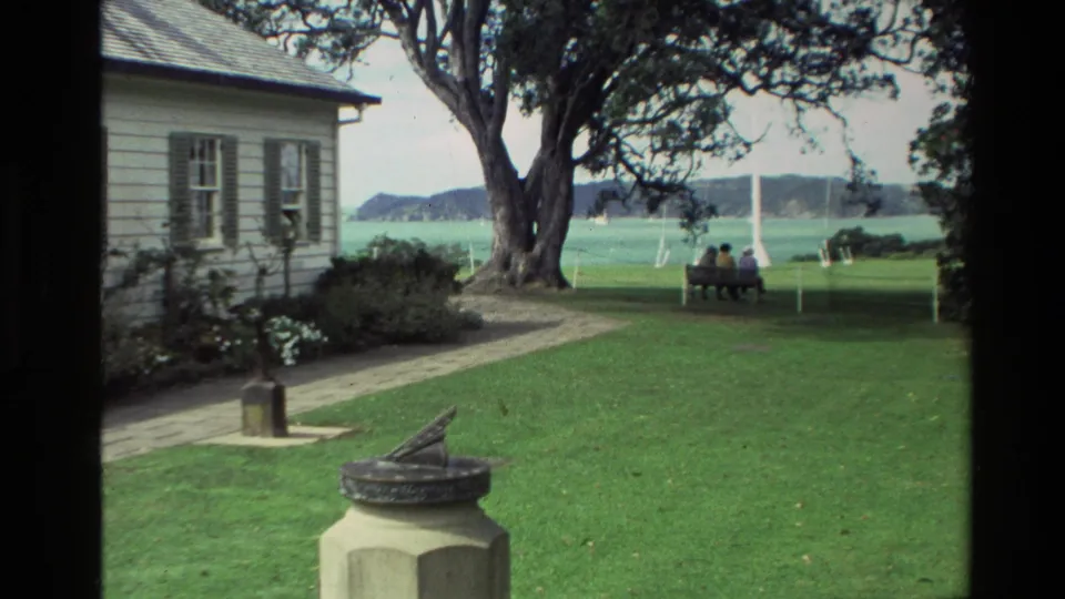 1981 New Zealand 8mm frame with a white house, garden path, sundial, large tree, waterfront, and two distant people seated on a bench.