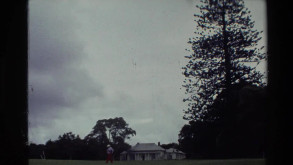 1981 New Zealand home-movie frame showing a man in a white shirt and red pants walking across a lawn near trees and a distant house.