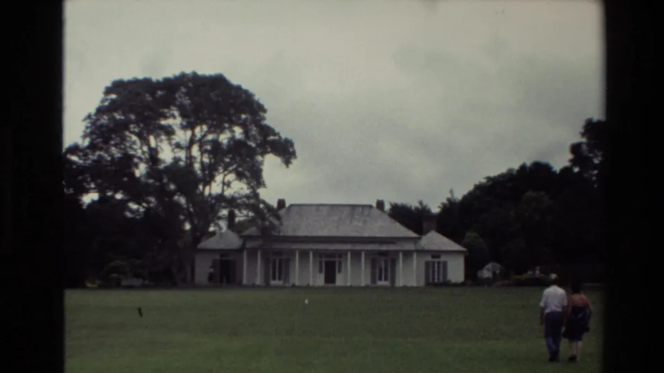 1981 New Zealand home-movie frame of two adults crossing a green lawn toward a large white house beneath mature trees.