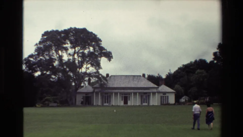 Wide 1981 New Zealand 8mm frame with two adults on a lawn approaching a large white house under cloudy skies.