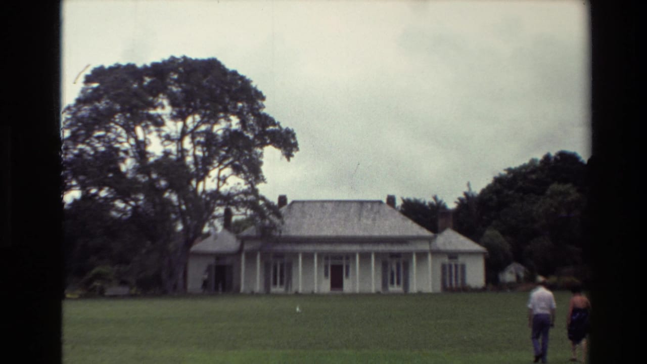 1981 New Zealand home-movie frame showing two adults walking across grass toward a white colonial-style house with a veranda and mature trees.