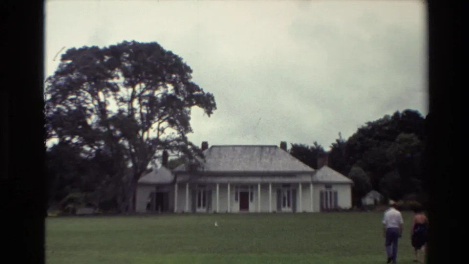 1981 New Zealand home-movie frame showing two adults walking across grass toward a white colonial-style house with a veranda and mature trees.