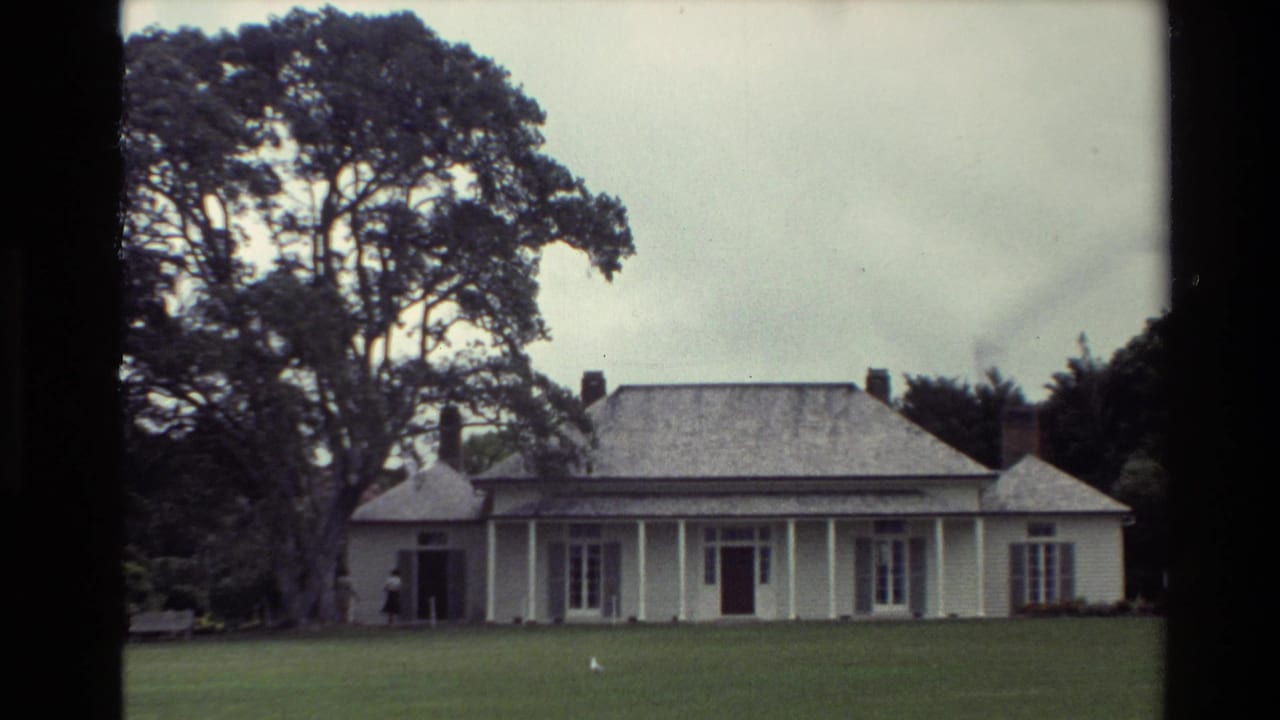 1981 New Zealand frame of a white weatherboard house with veranda columns, brick chimneys, broad lawn, and large trees under cloudy sky.