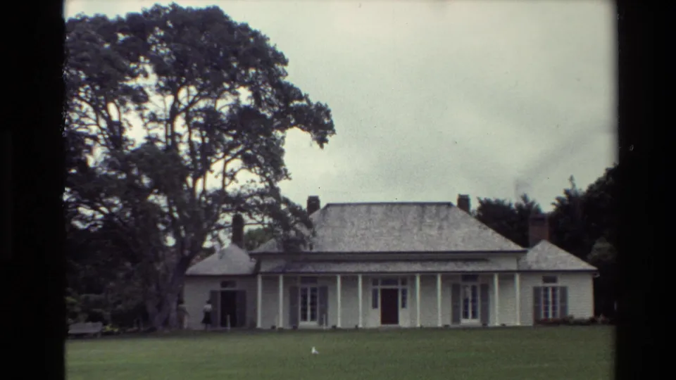 1981 New Zealand frame of a white weatherboard house with veranda columns, brick chimneys, broad lawn, and large trees under cloudy sky.
