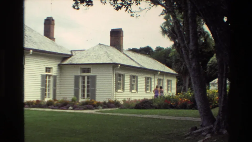 1981 New Zealand home-movie frame showing the side of a white house, tall windows, garden beds, lawn, trees, and adults near the flowers.