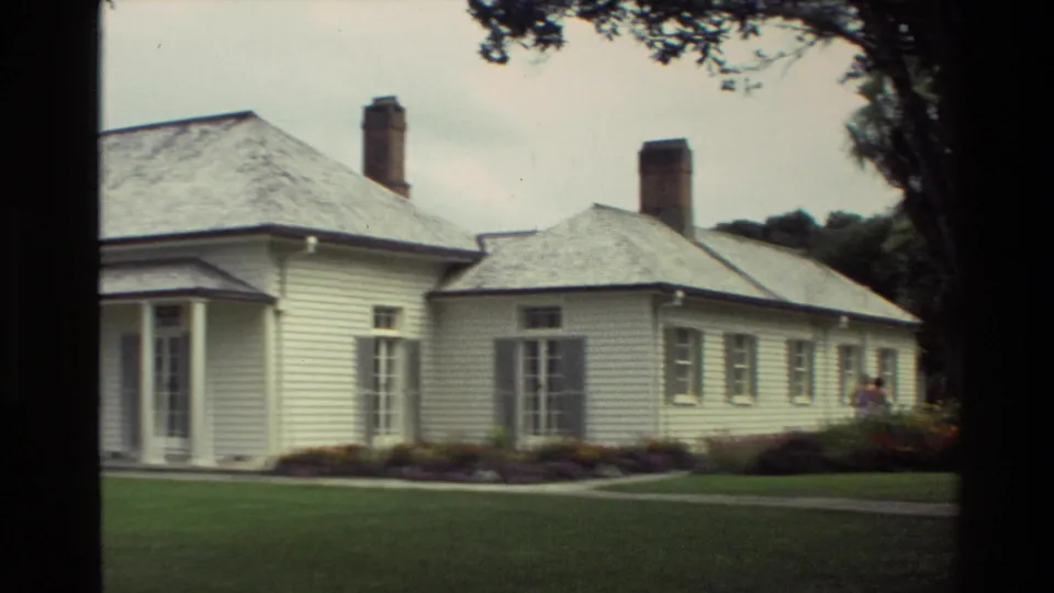 1981 New Zealand frame with a closer side view of the white house, brick chimneys, tall windows, garden beds, lawn, and adults partly visible at right.