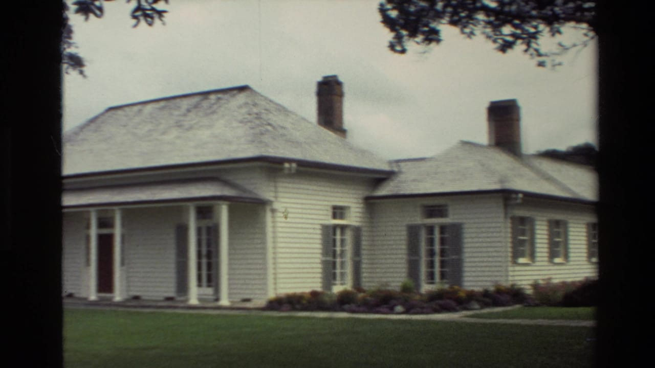 1981 New Zealand 8mm frame showing a white weatherboard house with a covered porch, brick chimney, lawn, garden beds, and overhanging trees.