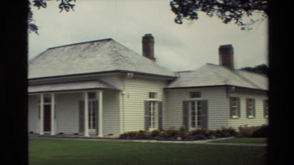 1981 New Zealand 8mm frame showing a white weatherboard house with a covered porch, brick chimney, lawn, garden beds, and overhanging trees.