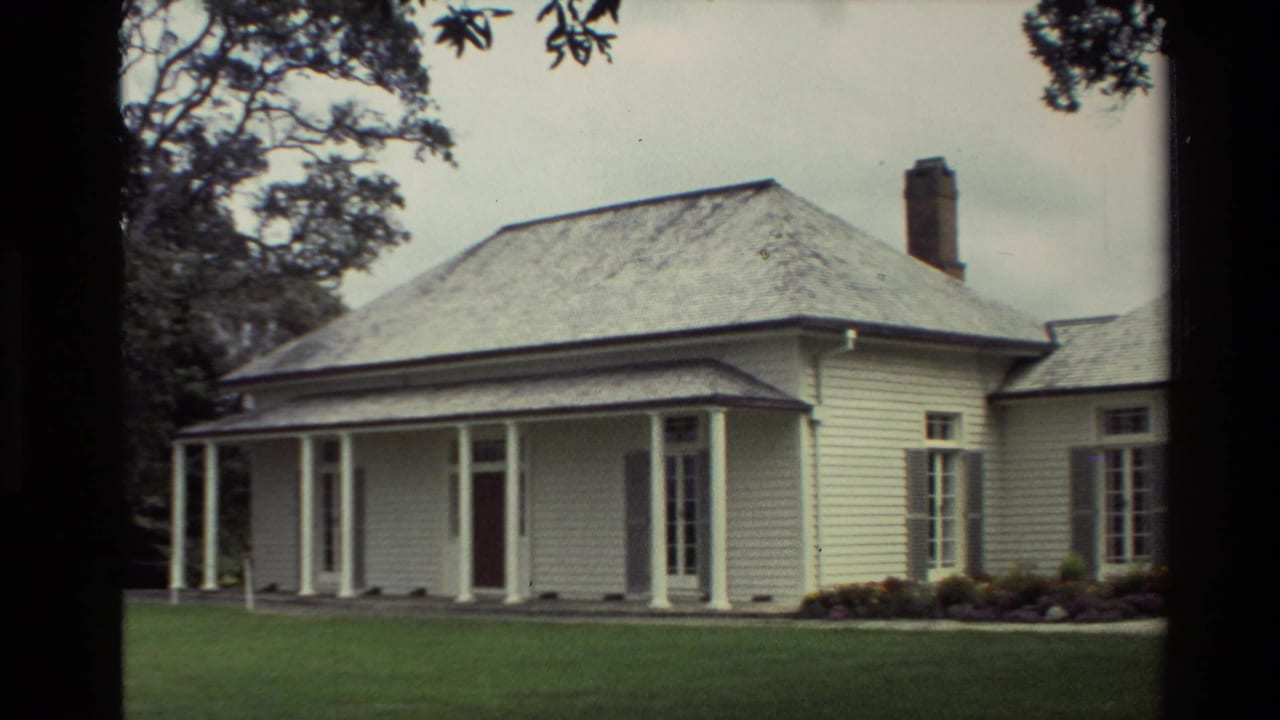 1981 New Zealand home-movie view of a white weatherboard house facade with porch columns, brick chimney, side wing, and lawn.