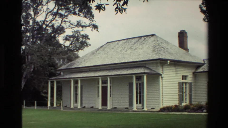 1981 New Zealand 8mm frame with a closer view of the white house porch, brick chimney, lawn, garden border, and tree trunk at left.