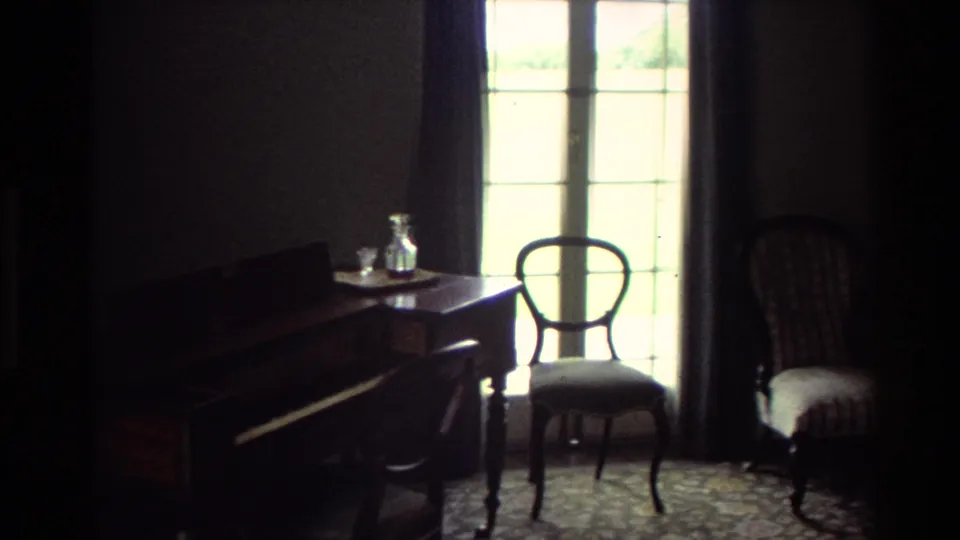 1981 New Zealand home-movie interior showing a dim room with piano, small table, glass decanter, chair, upholstered seat, and bright window.