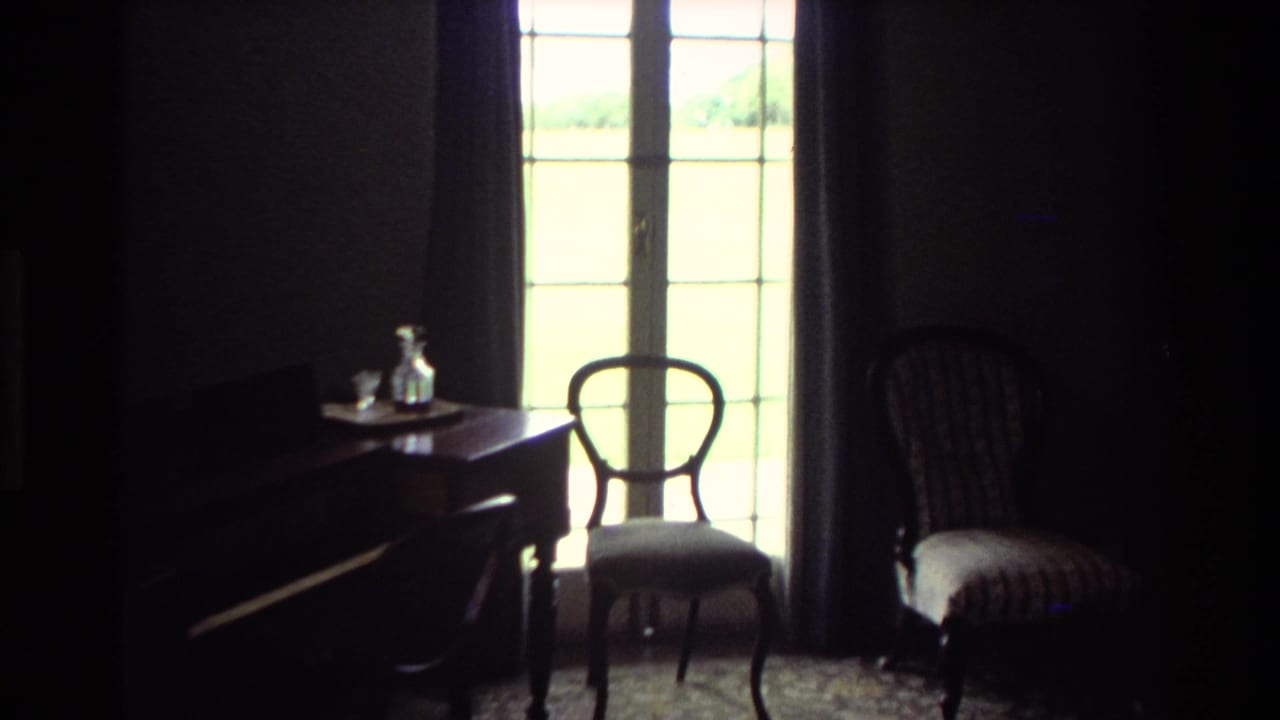 1981 New Zealand 8mm frame showing a dark room with French doors, wooden chairs, a desk and a small glass decanter.