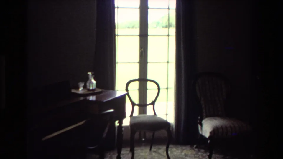 1981 New Zealand 8mm frame showing a dark room with French doors, wooden chairs, a desk and a small glass decanter.