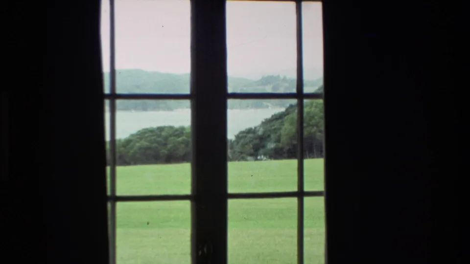 1981 New Zealand 8mm frame looking through French doors toward a green slope, wooded hills and a distant lake.