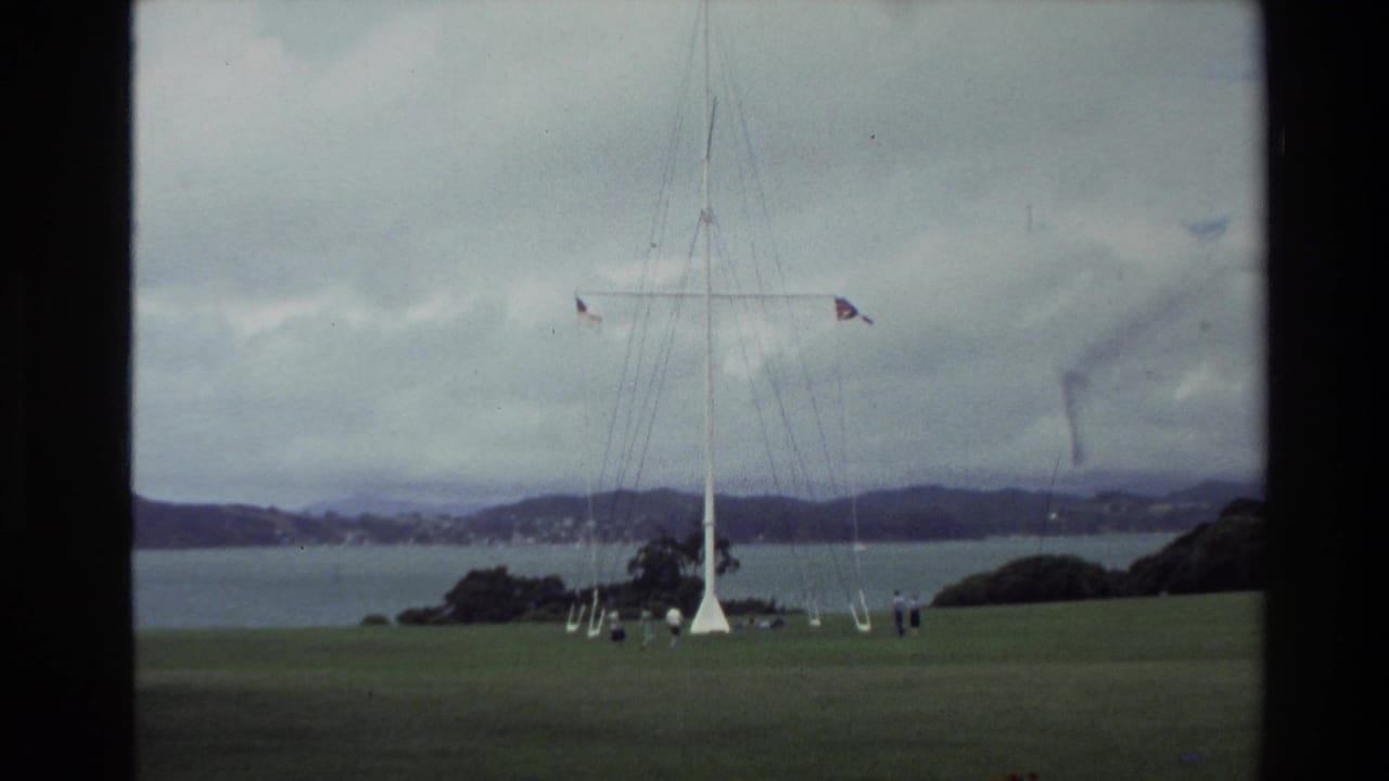 Tall flagpole on a grassy park above water, with small people nearby and hills in the distance.