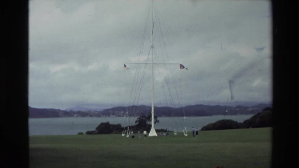 1981 New Zealand home-movie frame of a tall flagpole in a grassy waterfront park, with small visitors nearby.