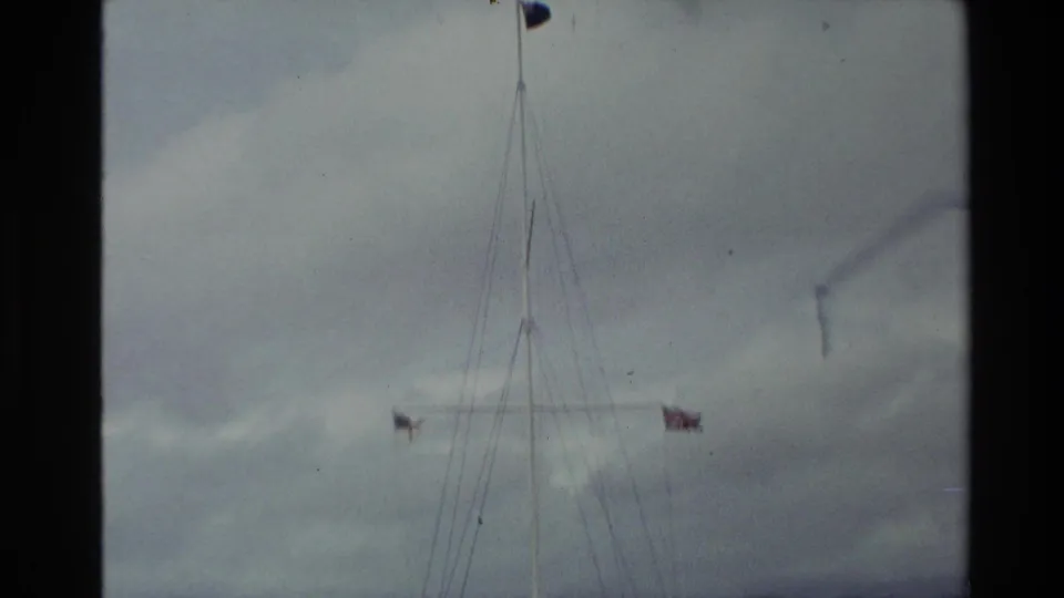 Close 1981 New Zealand 8mm view of the upper flagpole, rigging, and flags against a grey sky.