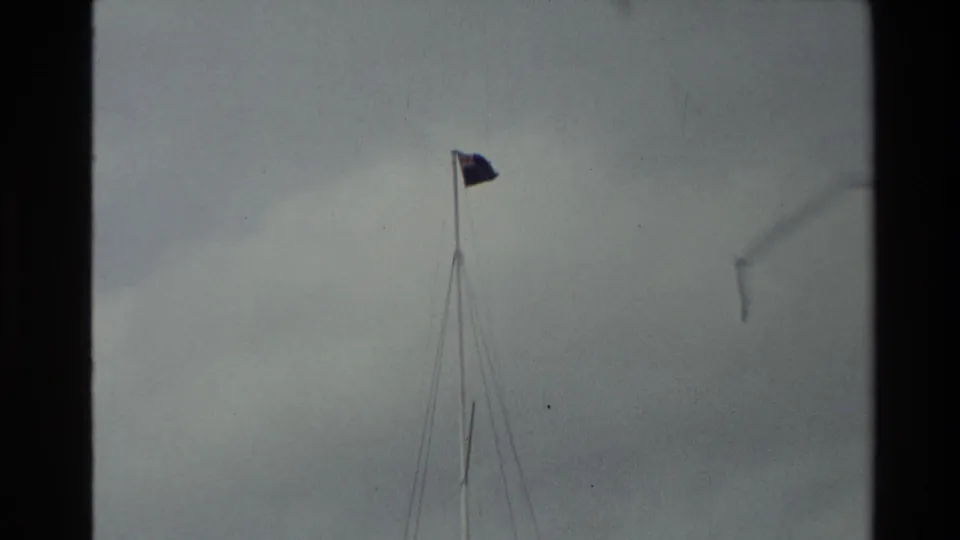 1981 New Zealand 8mm close view of the top of a tall flagpole, with a flag moving against overcast sky.