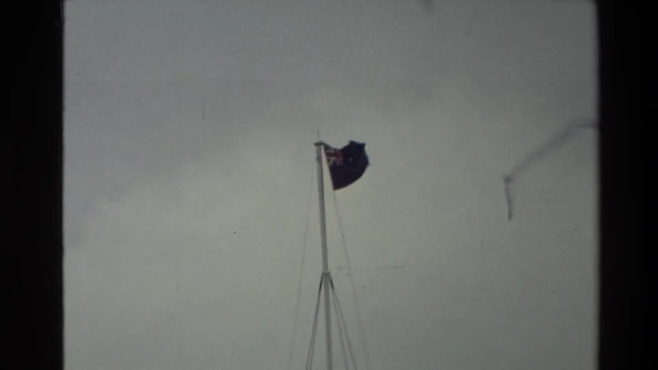 A wind-blown New Zealand flag appears near the top of a pole in a faded 8mm frame from the 1981 New Zealand clip.