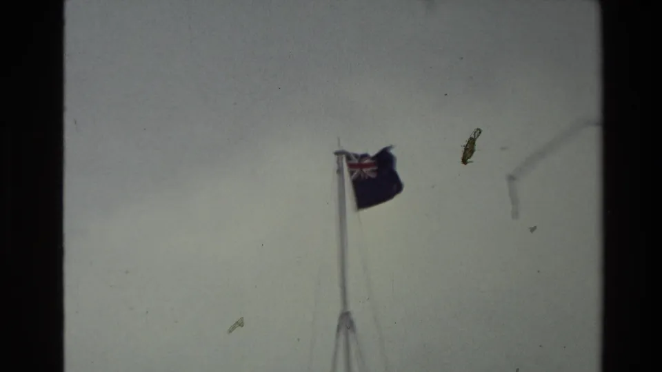 A closer 8mm view of the New Zealand flag and pole shows strong wind, pale sky, and visible film dust from the 1981 clip.